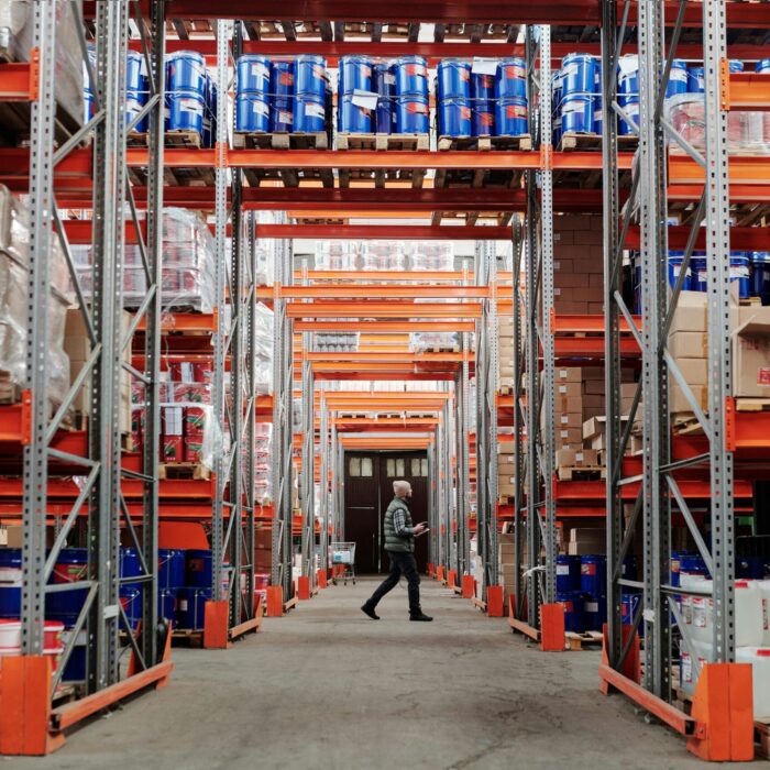 pexels-photo-4483775-4483775 A man walking through a large industrial warehouse with stacked shelves filled with goods and products.